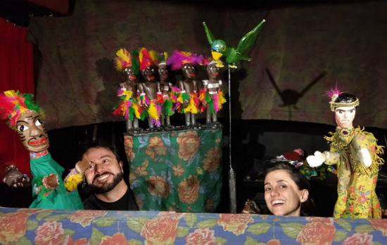 Foto de dois atores manipulando bonecos em cena de peça de teatro. Ao fundo, há 5 bonecos representando indígenas e um pássaro