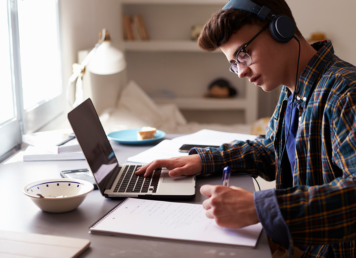 Foto de um adolescente de perfil escrevendo em um caderno e mexendo em um notebook. Ele usa óculos de grau e fone de ouvido (crédito: monkeybusinessimages – iStock)