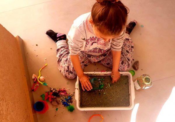 Foto vista de cima de uma menina sentada no chão brincando com uma caixa cheia de areia.