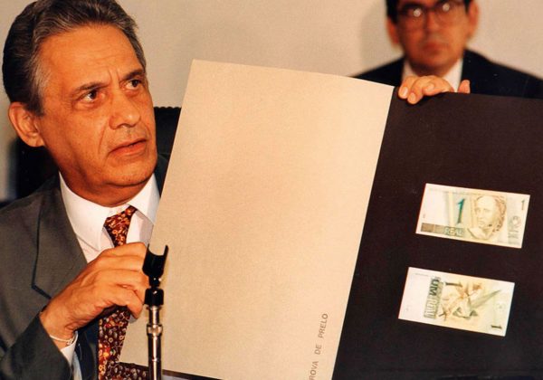 Foto, antiga, de Fernando Henrique Cardoso segurando uma pasta preta com uma duas notas de um real coladas nela, frente e verso.