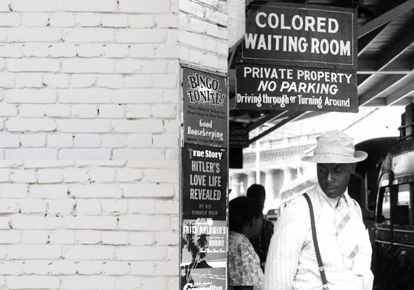 Foto antiga, em preto e branco, de um homem negro parado em uma estação de trem. Sobre ele, há uma placa com o texto "colored waiting room".