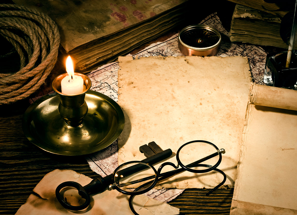 Foto em detalhe de uma mesa antiga, Sobre ela, há papéis amarelados, uma vela, uma chave e um óculos antigo (ulkan – iStock)