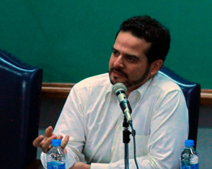 Foto do professor Flávio Carvalho. Ele usa camisa branca e discursa em um microfone enquanto gesticula com as mãos