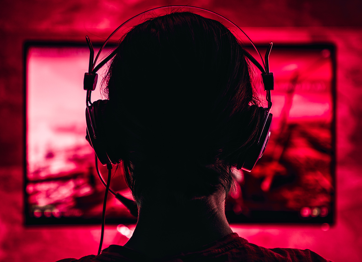 Foto em detalhe, contra a luz e tirada de trás da cabeça de uma pessoa com fone de ouvido gamer. Atrás dela, há um monitor de computador