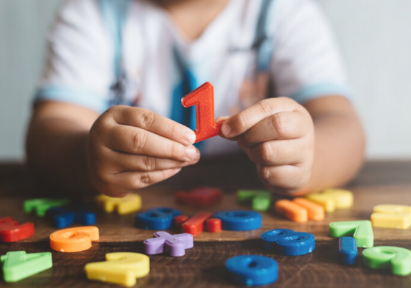 Foto em close das mãos de uma criança segurando o número um, abaixo há o topo de uma mesa com vários outros números de plásticos dispersos.