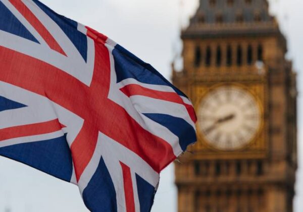 Bandeira do Reino Unido (Union Jack) em destaque, com a torre do relógio Big Ben desfocada ao fundo em Londres.