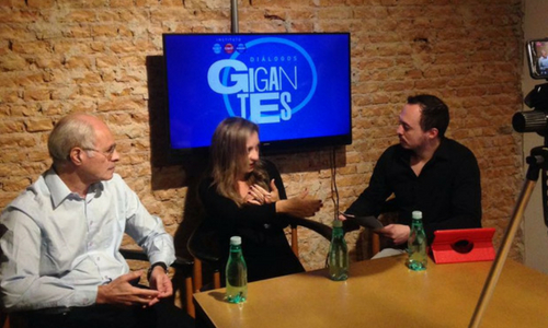 Foto de três pessoas durante um debate. Ao fundo, há uma tela com o título Diálogos Gigantes