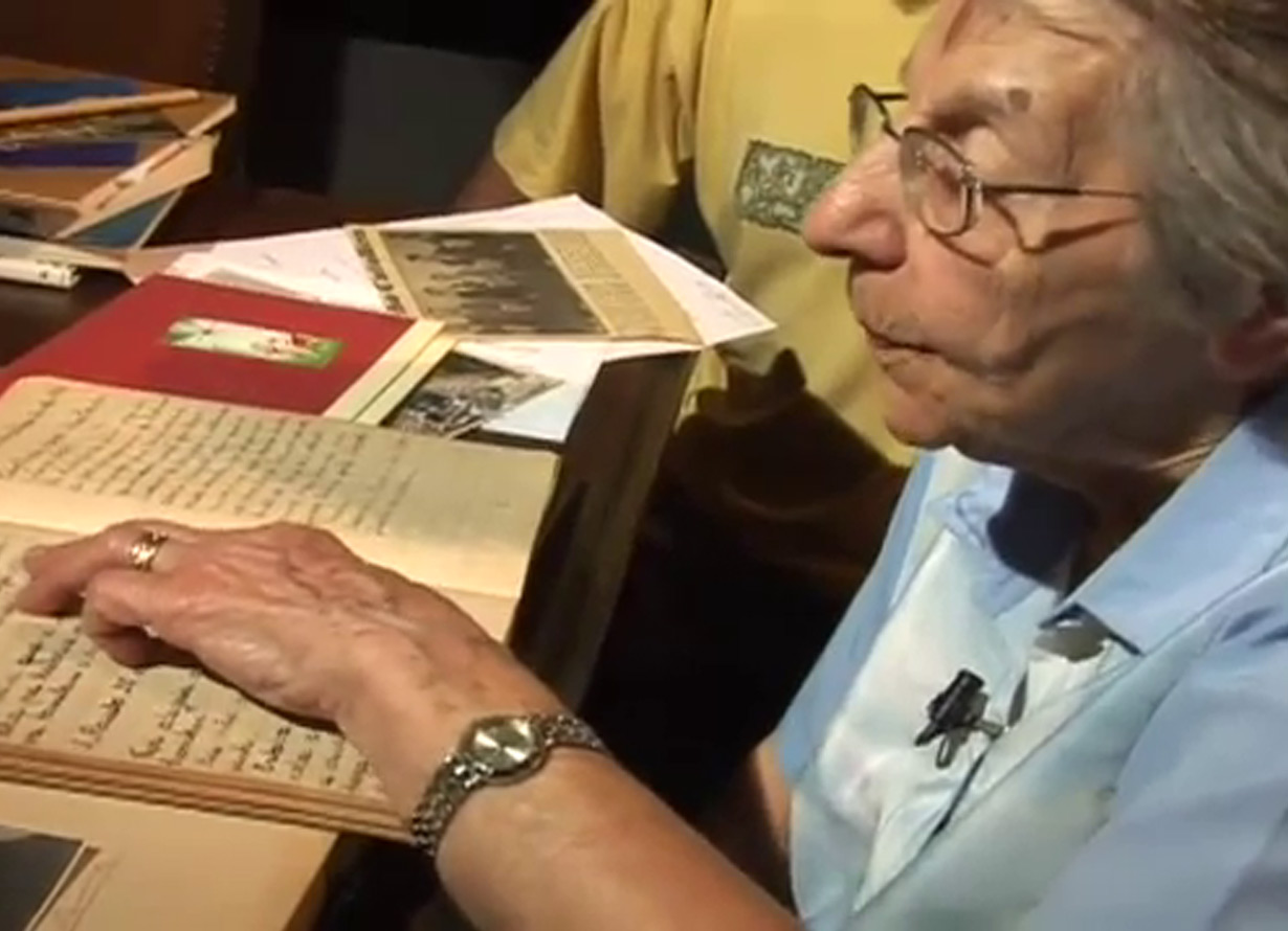 Foto de uma idosa de perfil. Ela passa o dedo em um diário antigo com as páginas por textos