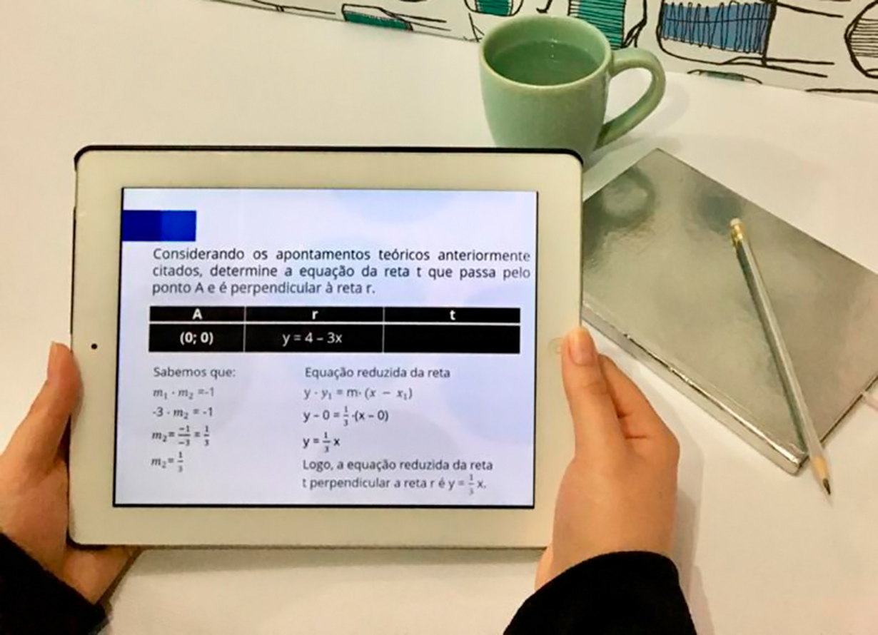 Foto de um par de mãos segurando um tablet, na tela deste há uma questão matemática. Ao fundo, sobre uma mesa, há uma caneca, caderno e lápis.
