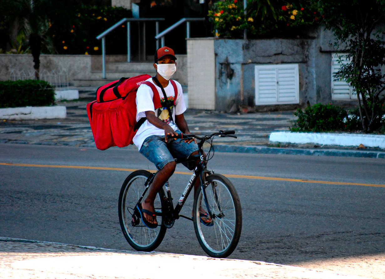 Foto de um homem de boné e chinelo montado em uma bicicleta. Ele usa máscara de tecido no rosto e uma bolsa de entregas de comida nas costas
